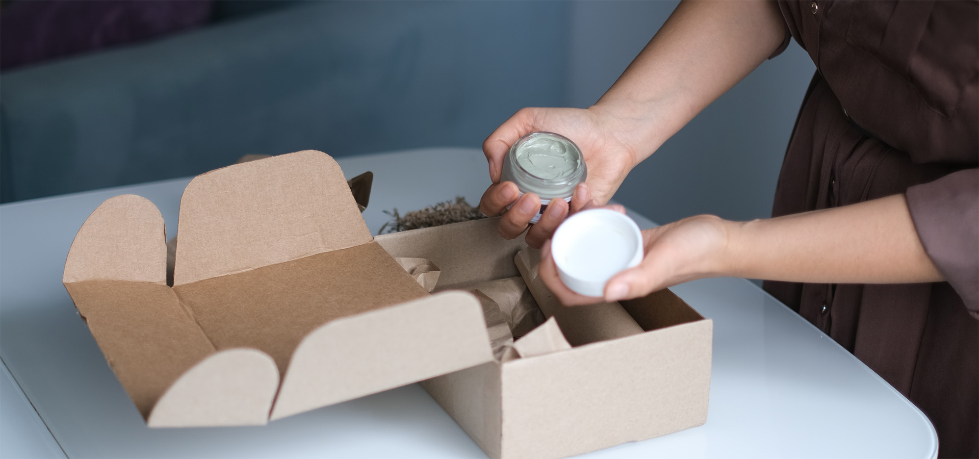 Hände halten ein Kosmetikprodukt und dessen Deckel, während ein Karton für die Verpackung vorbereitet wird – Beispiel für die operative Umsetzung von Marketing- und Promotionsaktionen bei Greim Projekt Service.