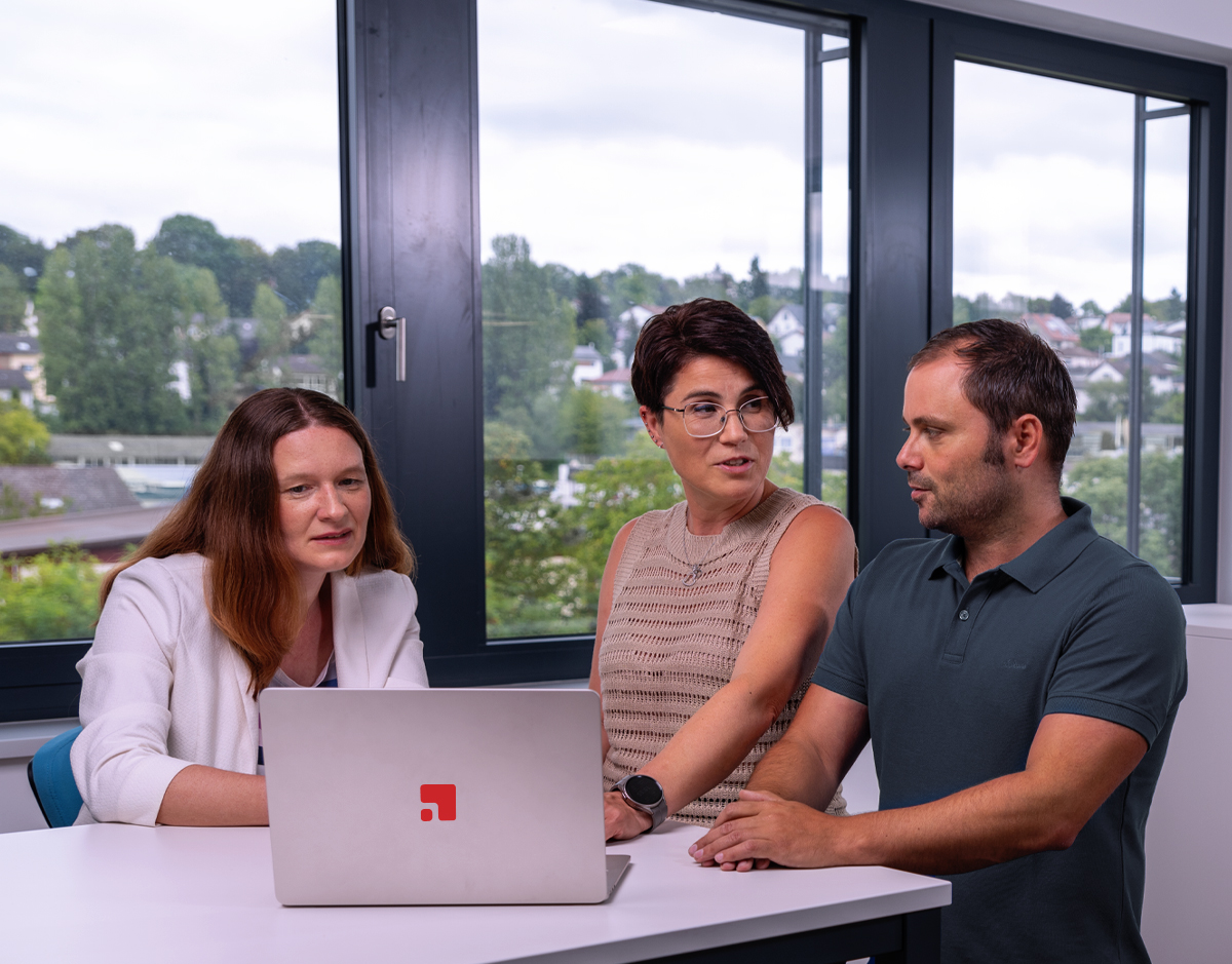 Drei Mitarbeiter von Die Greim Projekt Service GmbH arbeiten gemeinsam an einem Laptop in einem modernen Büro mit Blick auf Grünflächen und Häuser – sie planen oder umsetzen Marketing- oder Kampagnenprojekte, wie das Unternehmen für organisatorische und operative Dienstleistungen spezialisiert ist.