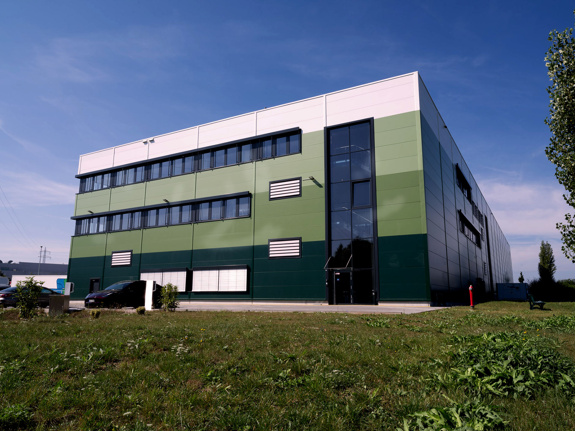 Modernes Bürogebäude der Greim Projekt Service GmbH in Wörrstadt mit grün-weißer Fassade, Glasfront und Parkplatz, umgeben von Grünfläche.