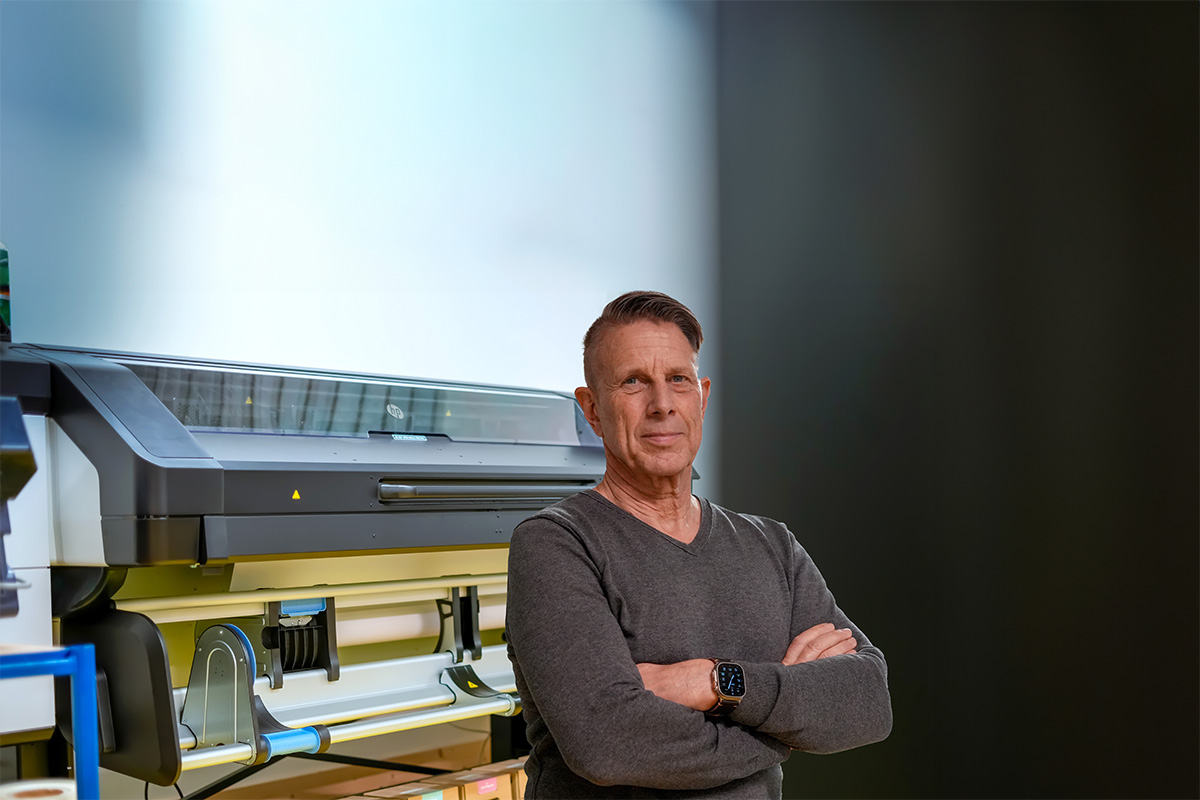 Jochen Greim von Greim Projekt Service steht vor einer großen industriellen Druckmaschine, die für die operative Umsetzung von Marketing- und Promotionsprojekten genutzt wird.