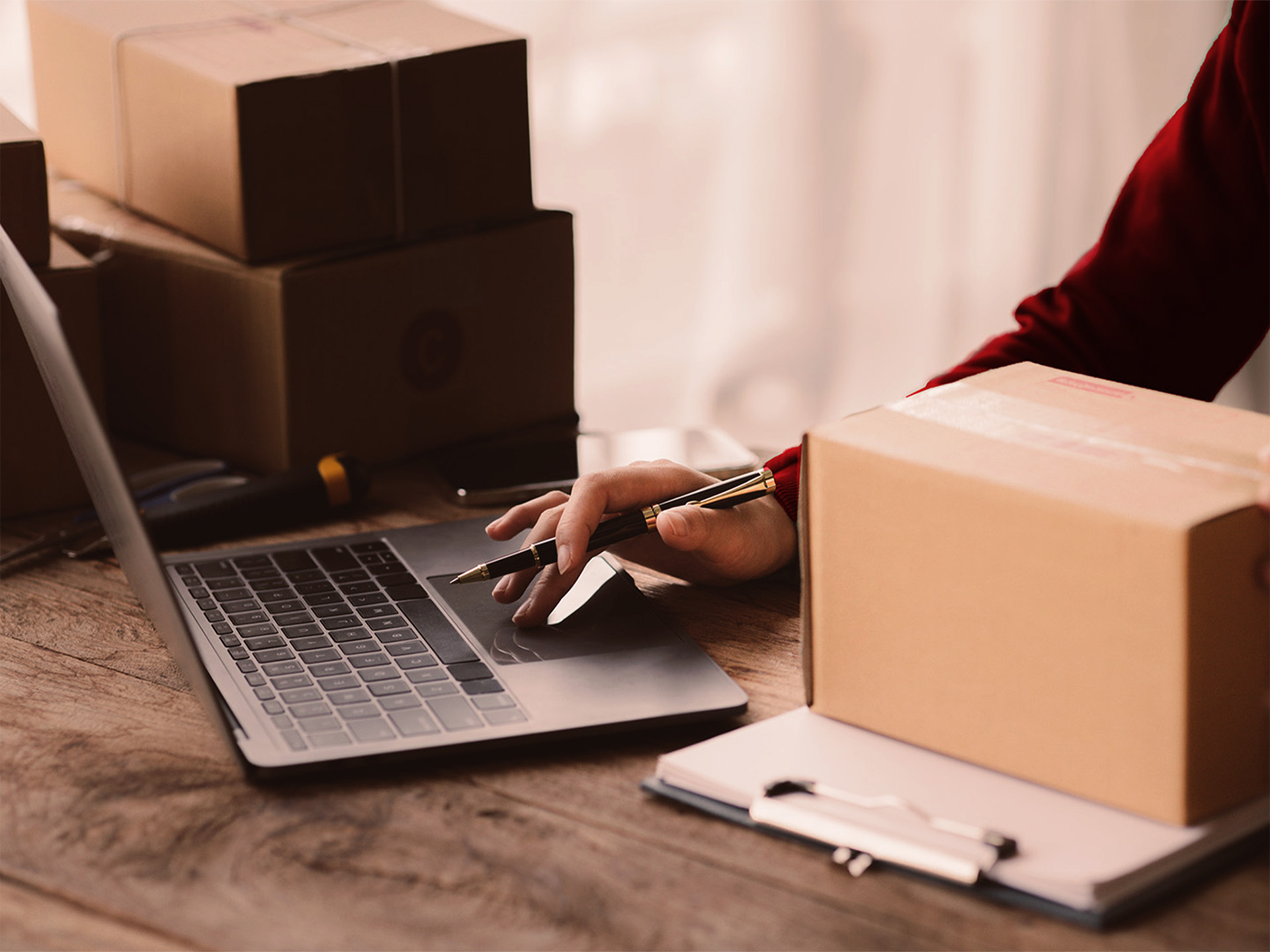 Hand mit Stift am Laptop, umgeben von Versandkartons und einem Clipboard, bei der organisatorischen oder logistischen Abwicklung von Marketing- oder Promotionsprojekten.