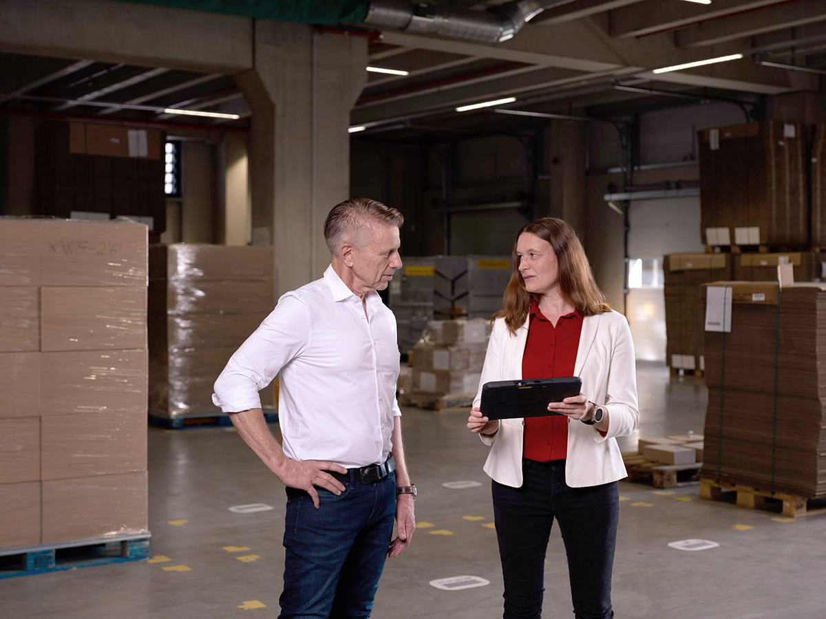 Jochen Greim und Christina Richter von Greim Projekt Service GmbH diskutieren in einem Lager über logistische Prozesse oder Marketingprojekte, wobei eine Person ein Tablet zur Präsentation verwendet.