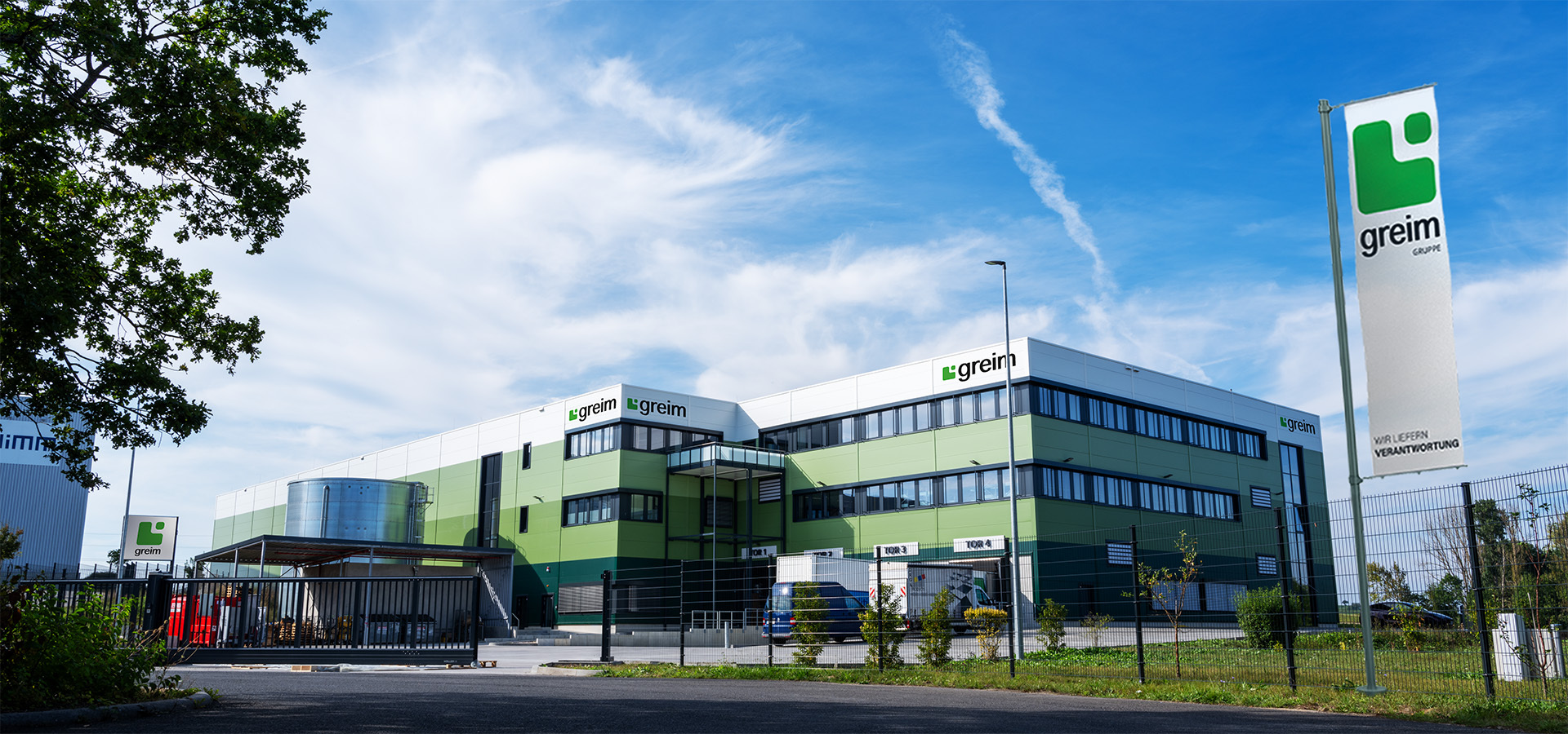 Modernes Bürogebäude der Greim Projekt Service GmbH mit grün-weißer Fassade und greim-Logos, umgeben von einem Zaun und Fahrzeugen – Sitz des Dienstleistungsunternehmens für Marketing-, Promotions- und Kampagnenprojekte in Wörrstadt.