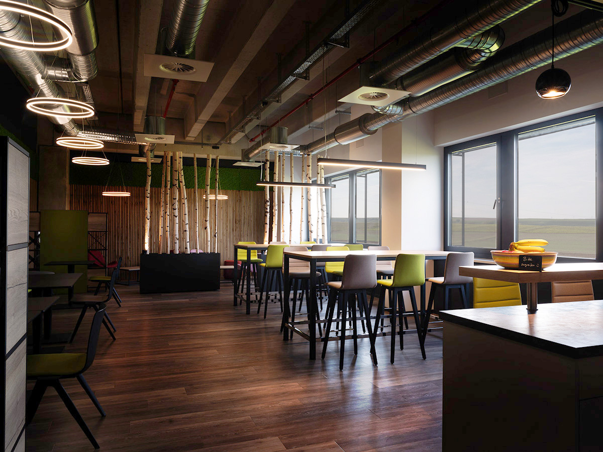 Modernes Lounge- und Aufenthaltsraum mit industrieller Design (exponierte Rohre, Holzparkett), großen Fenstern, bunten Sitzmöbeln und Birkenstamm-Dekoration – ein professioneller und einladender Raum für Pausen oder informelle Meetings.
